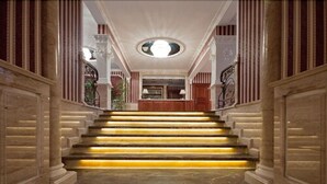 Una imponente escalera de mármol con candelabro, que conduce a una sala con chimenea y puerta de madera.