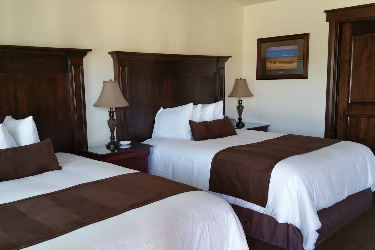 A hotel room with two beds, wooden headboards, white bedding, brown throw pillows, bedside lamps, a nightstand, and a framed picture on the wall.