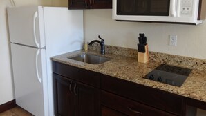 A kitchen with a white refrigerator, a microwave, a granite countertop, a sink, and dark wooden cabinets. - Winterton Suites (Williston)
