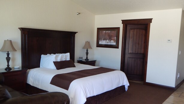 A bedroom with a large bed, a wooden headboard, two bedside tables with lamps, a brown leather chair, a ceiling fan, a door, and a framed picture on the wall. - Winterton Suites (Williston)