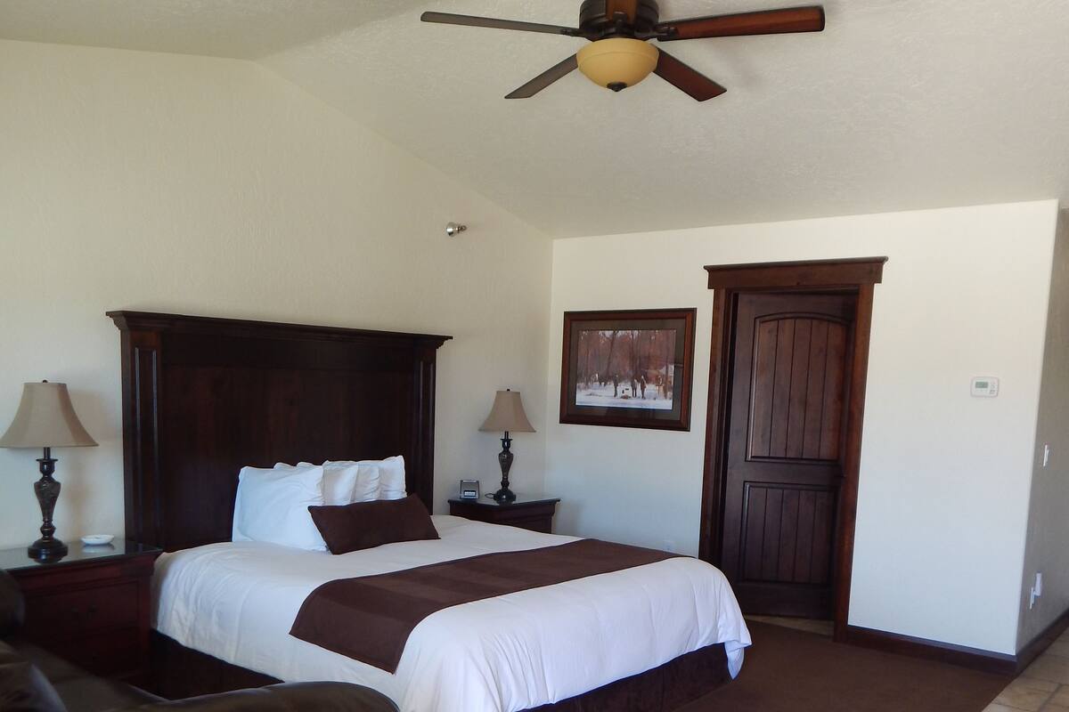 A bedroom with a large bed, a wooden headboard, two bedside tables with lamps, a brown leather chair, a ceiling fan, a door, and a framed picture on the wall.