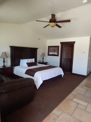 A bedroom with a large bed, a wooden headboard, two bedside tables with lamps, a brown leather chair, a ceiling fan, a door, and a framed picture on the wall. - Winterton Suites (Williston)
