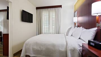 Una habitación de hotel con una cama grande, una mesita de noche con lámpara, un televisor montado en la pared y una ventana con cortinas translúcidas.