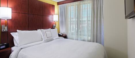 A hotel room with a large bed, two bedside tables with lamps, a TV mounted on the wall, and a window with sheer curtains.
