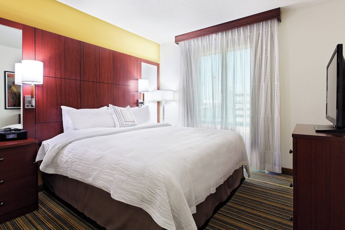 A hotel room with a large bed, a wooden headboard, a flat-screen TV, and a window with sheer curtains.