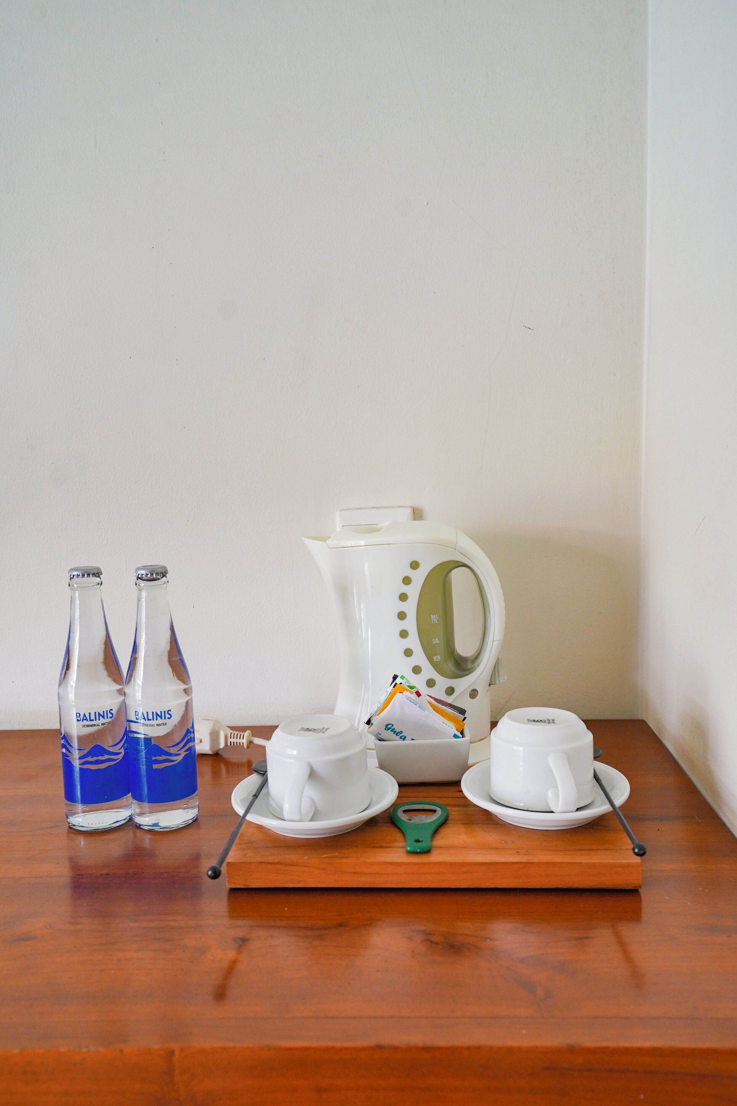 A table with two bottles of water, a white electric kettle, two white cups, and a green bottle opener on a wooden tray.