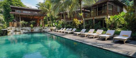 A pool area with lounge chairs and a wooden deck, surrounded by lush greenery and palm trees.