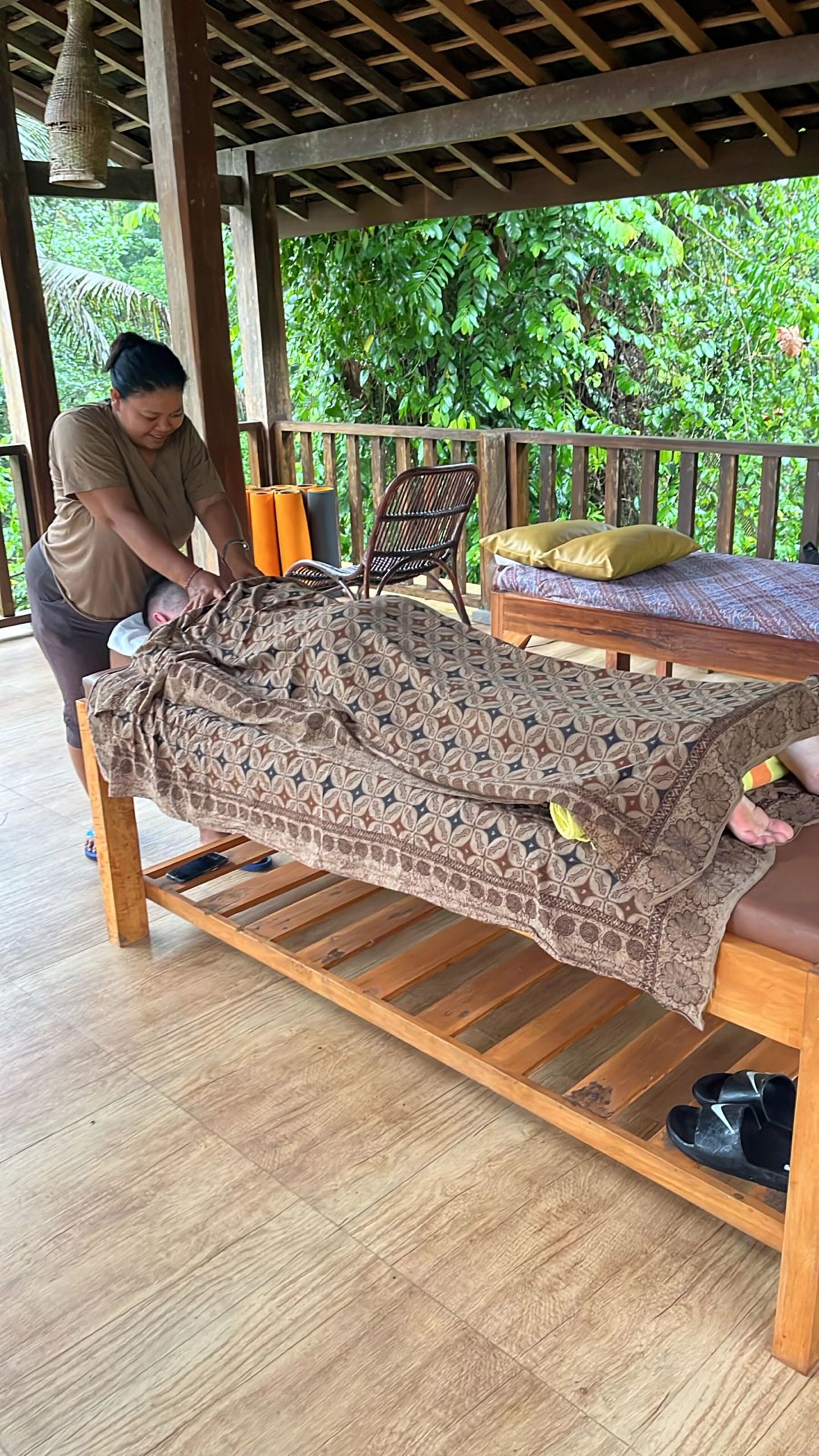 Een houten bed met een bedsprei met patroon, een houten stoel en een houten tafel met kussens.