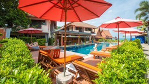 A poolside area with lounge chairs and red umbrellas, surrounded by greenery and a building with a pool. - Sunrise Resort (Ko Pha-ngan)