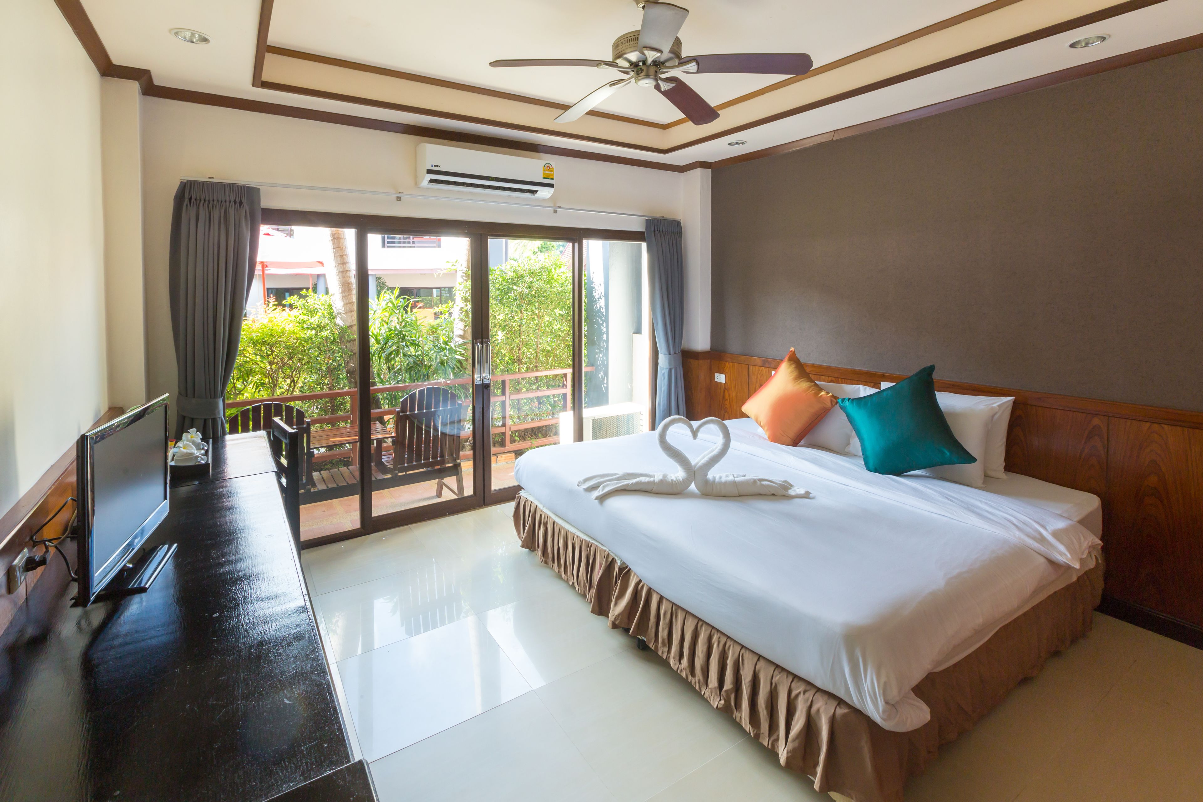 Habitación de hotel con cama, escritorio con computadora, ventilador de techo y puerta corrediza de vidrio al exterior.