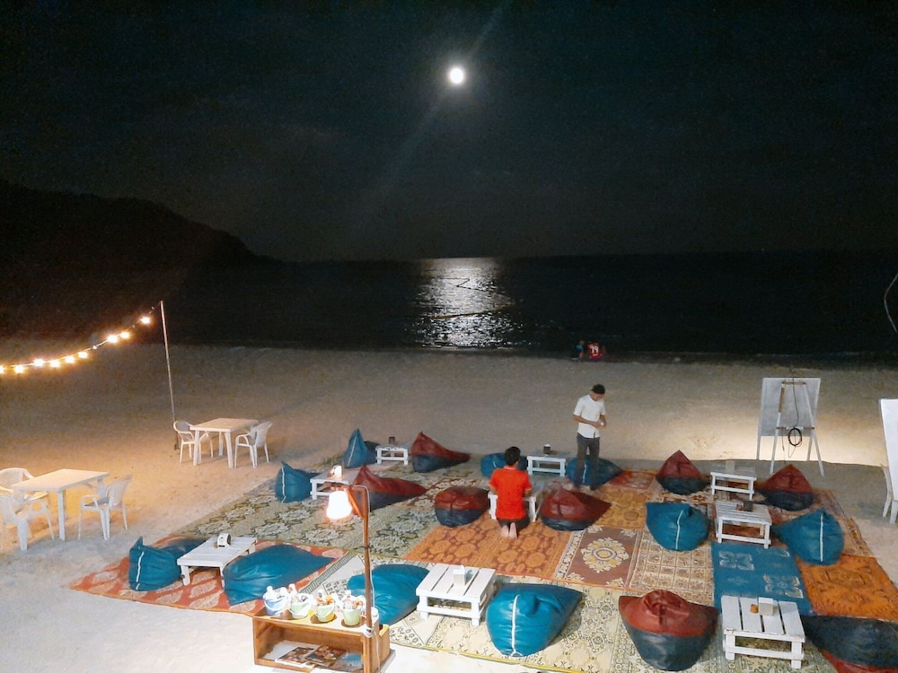 A beachside seating area with tables and chairs, bean bags, and a small table with drinks.