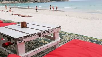 A beach with a white table and red cushions, a rocky hillside in the background, and people walking on the sand.