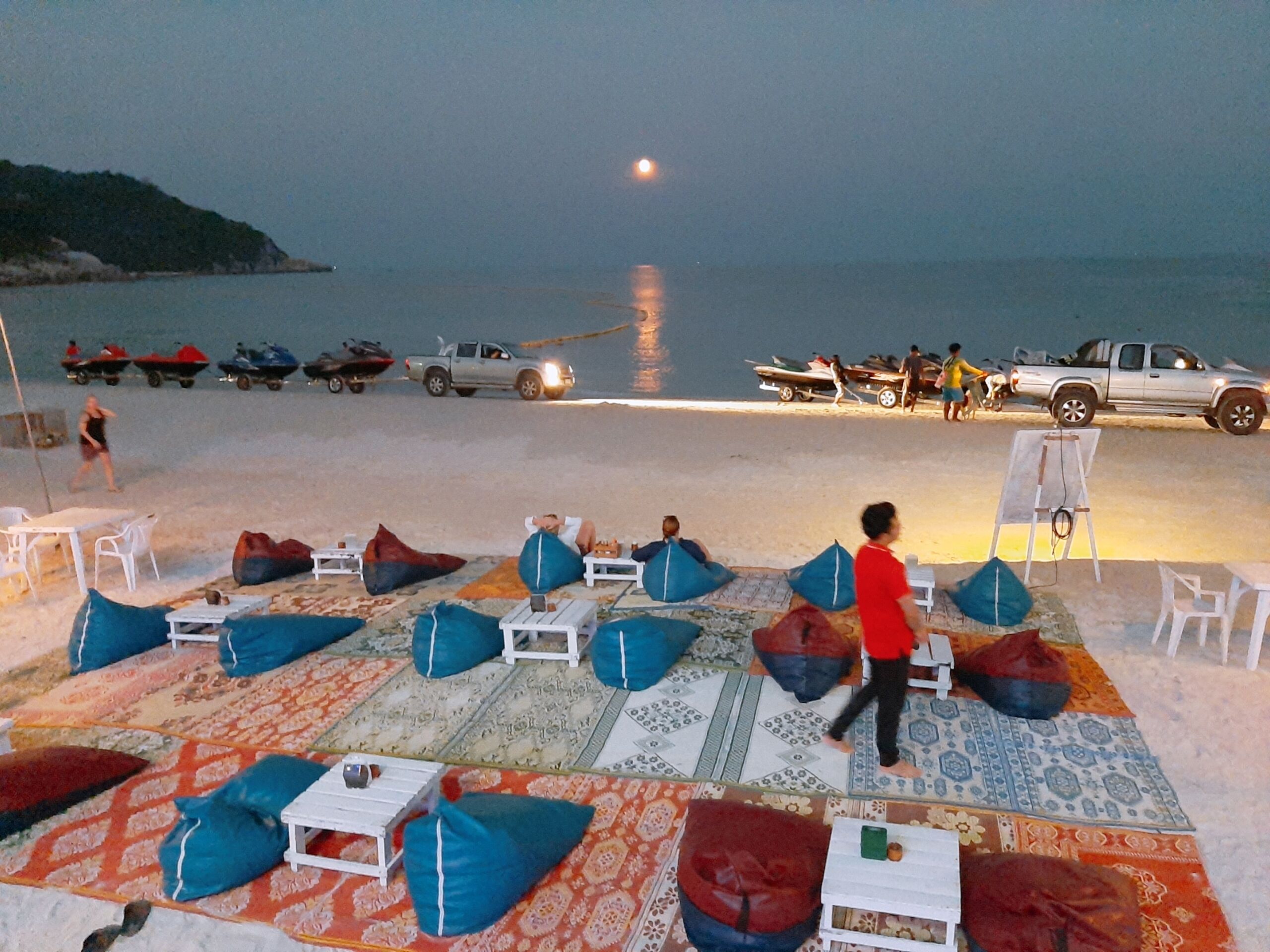 Un espace aménagé au bord de la plage, avec des chaises longues et des fauteuils en forme de sac, une petite scène et une vue sur la mer et les bateaux.