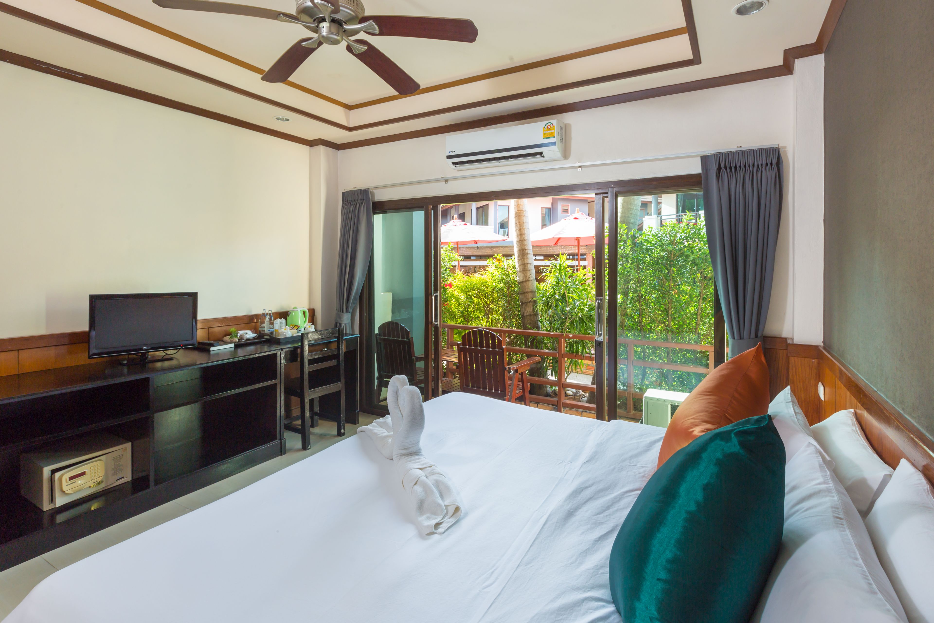 A hotel room with a bed, a desk with a television, and a balcony with a view of greenery and buildings.