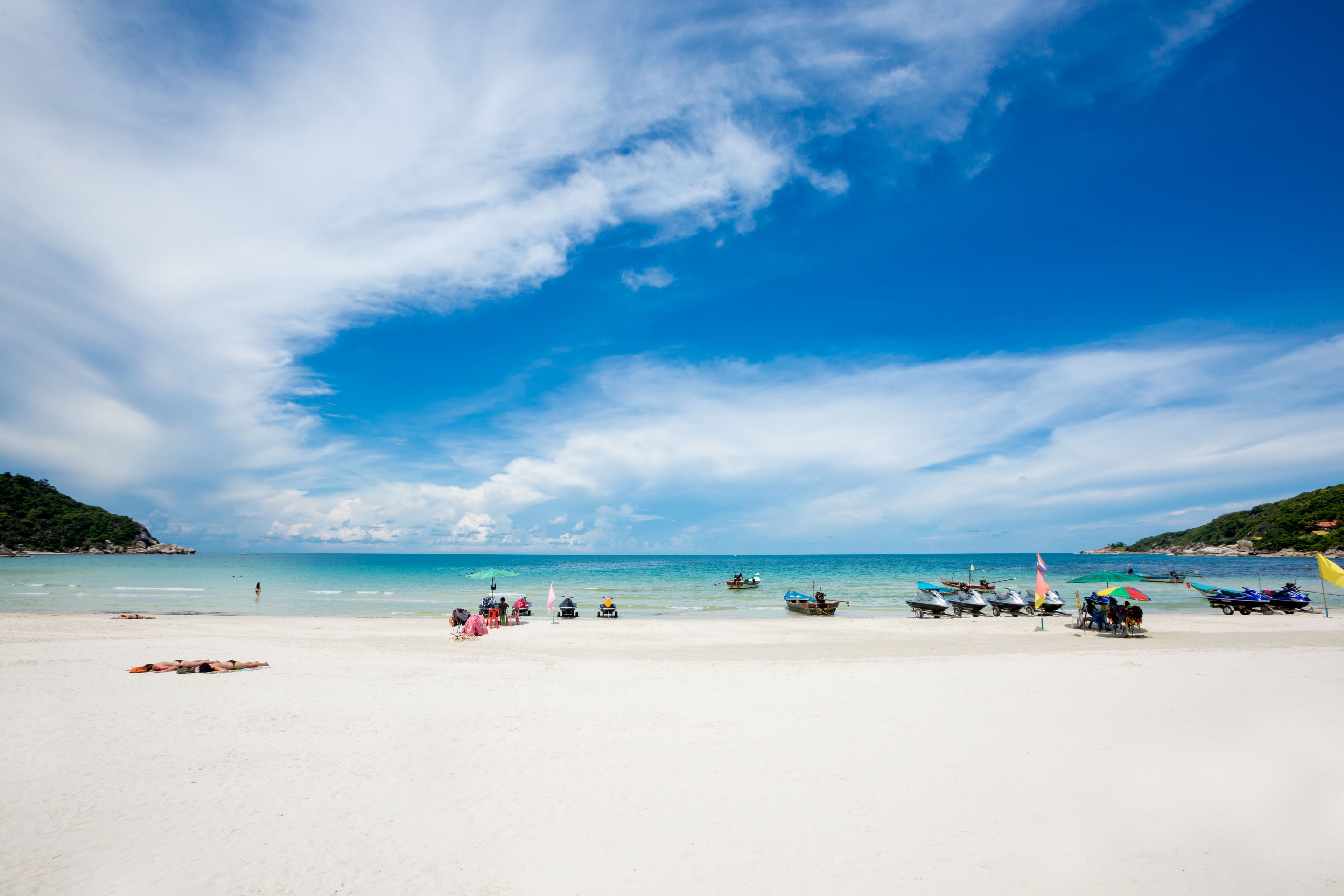 A beach with white sand, clear blue water, and boats.