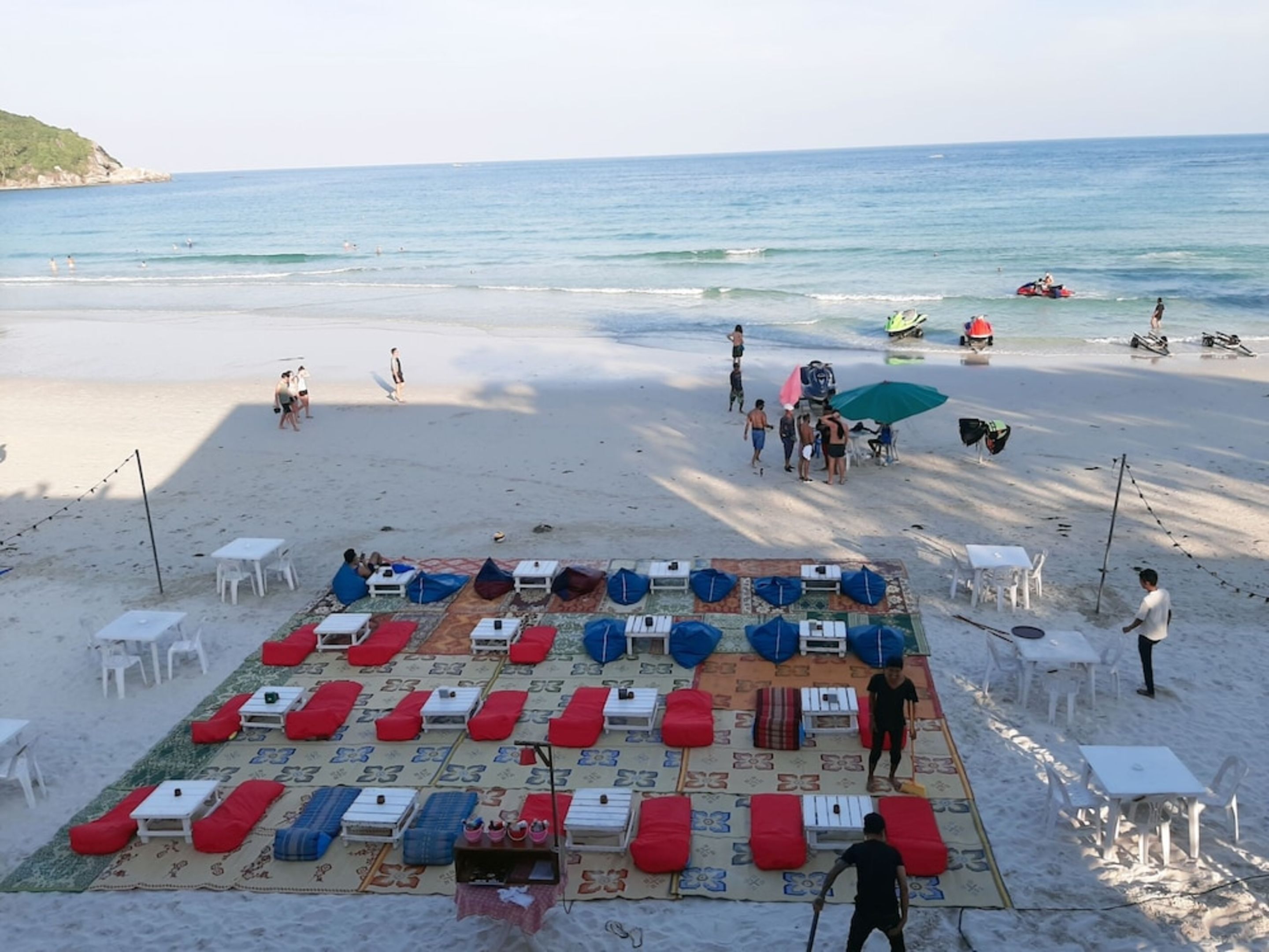 A beach with sun loungers and tables arranged on a mat, a few people walking, and boats in the water.