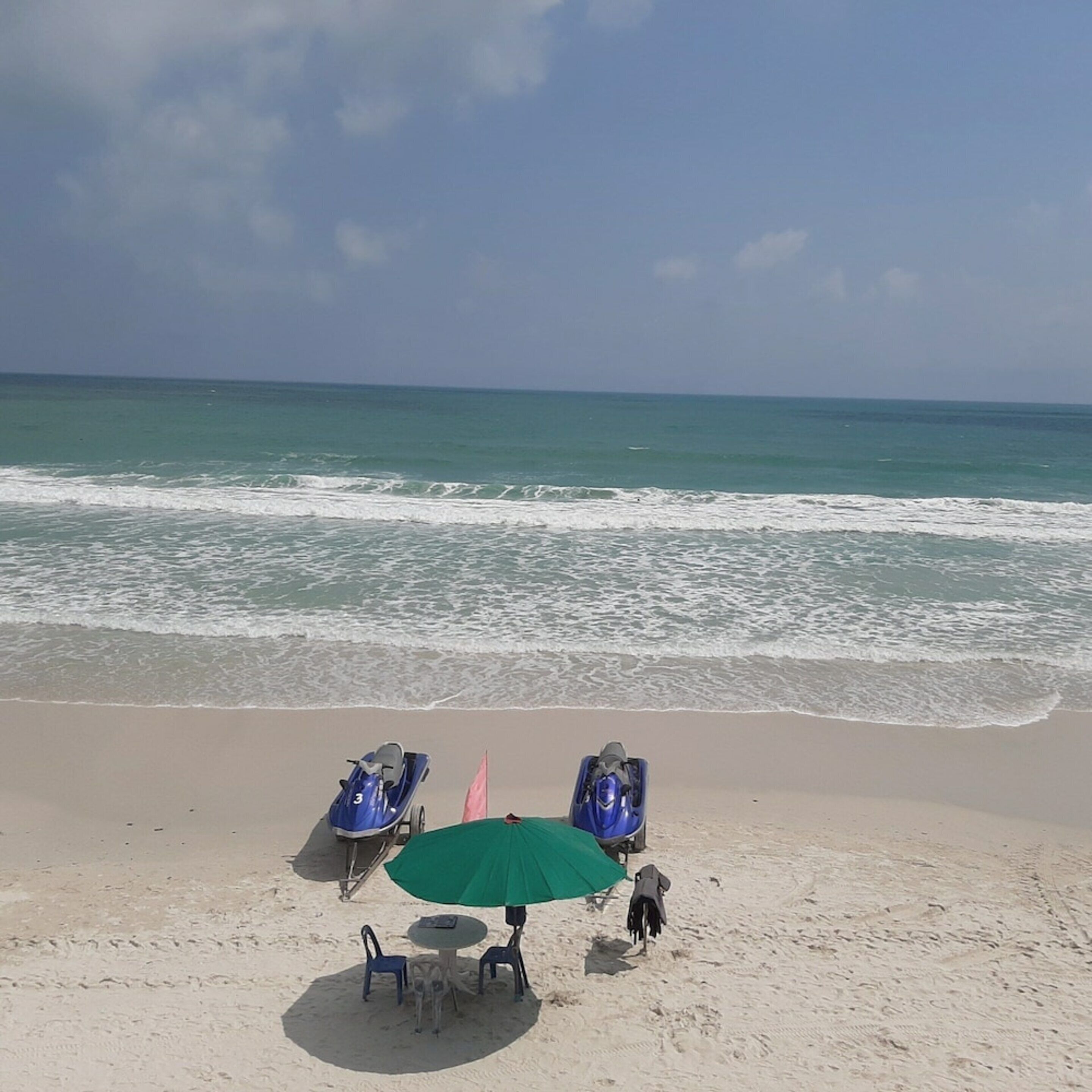 A beach with a green umbrella, two blue chairs, and a jet ski.