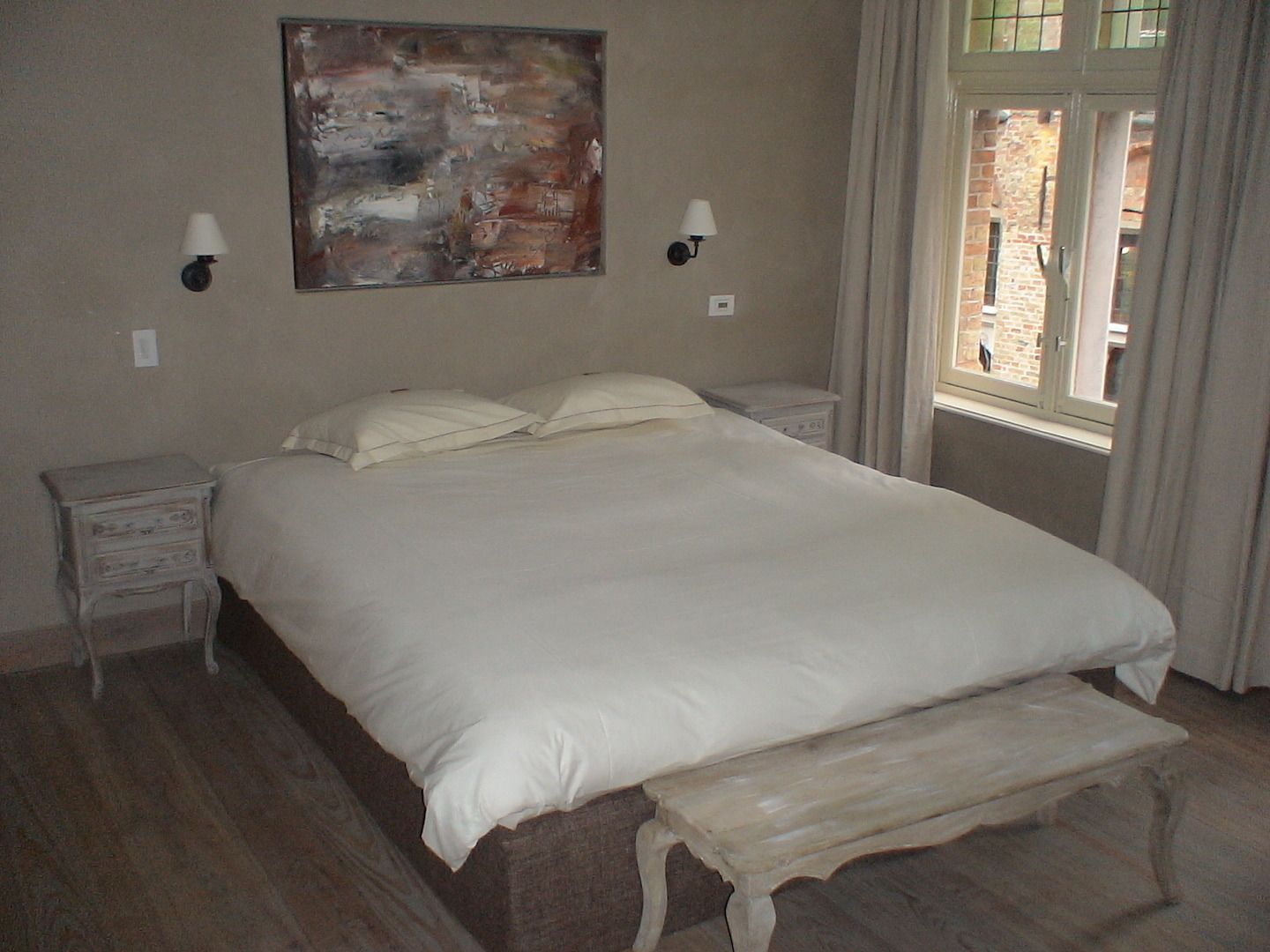 Une chambre à coucher avec un grand lit, deux tables de chevet, un banc, un tableau et une fenêtre avec des rideaux.