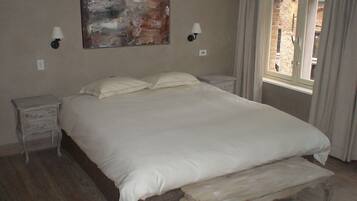 Une chambre à coucher avec un grand lit, deux tables de chevet, un banc, un tableau et une fenêtre avec des rideaux.