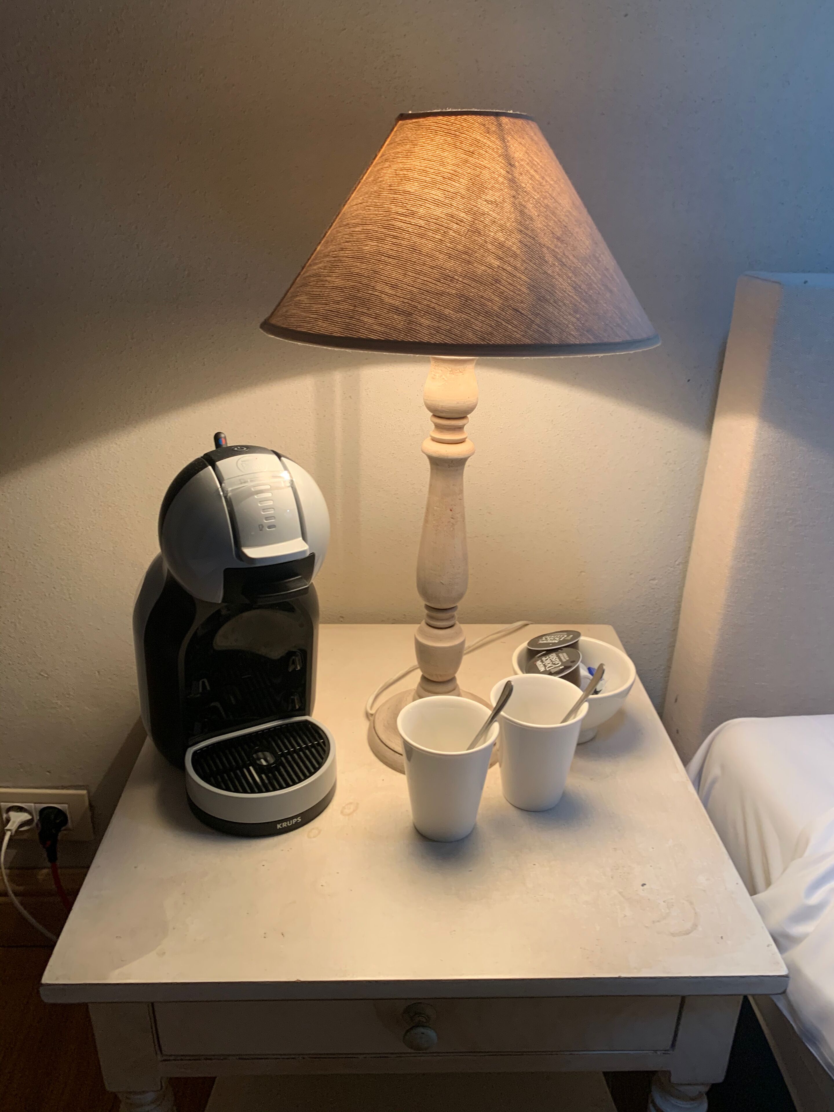 Ein weißer Nachttisch mit einer Kaffeemaschine, einer Lampe und zwei weißen Tassen.
