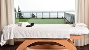 A room with a massage table, towels, a small black bowl, and a glass railing overlooking a green area and a body of water.