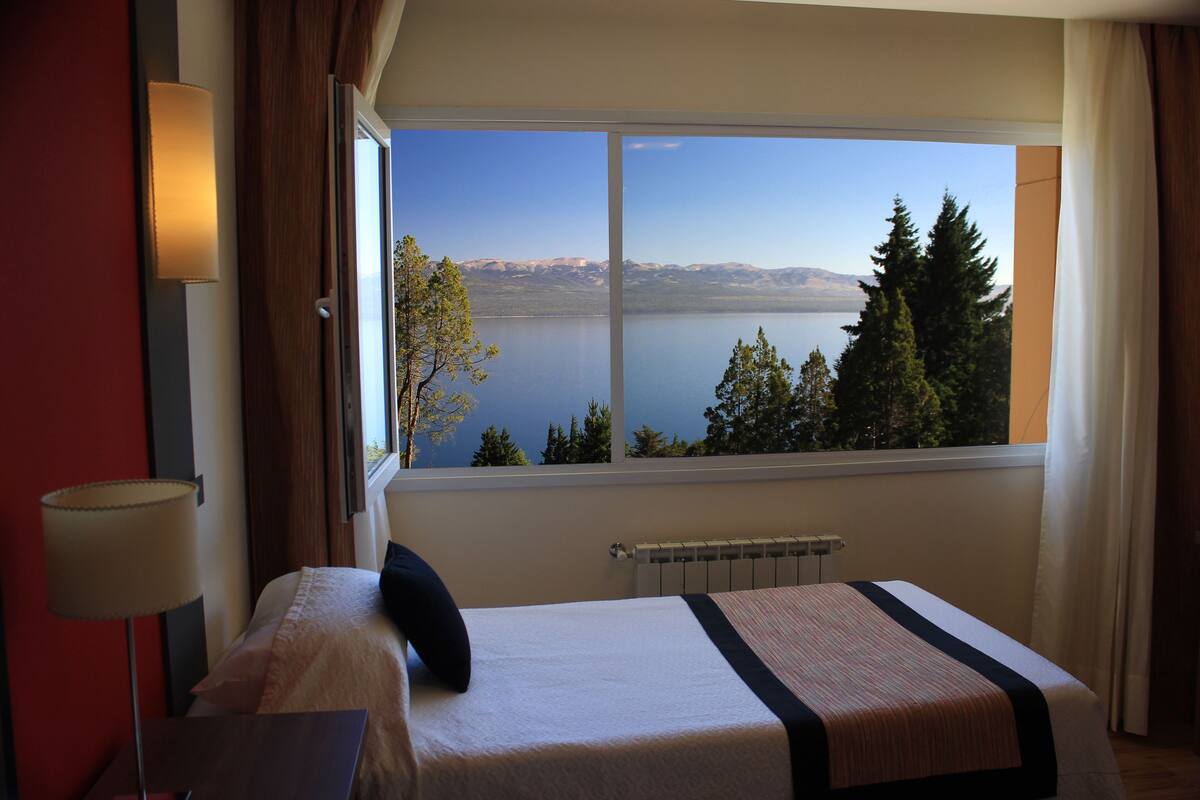 Ein Hotelzimmer mit großem Fenster, einem Bett, einem Nachttisch und einer Lampe.
