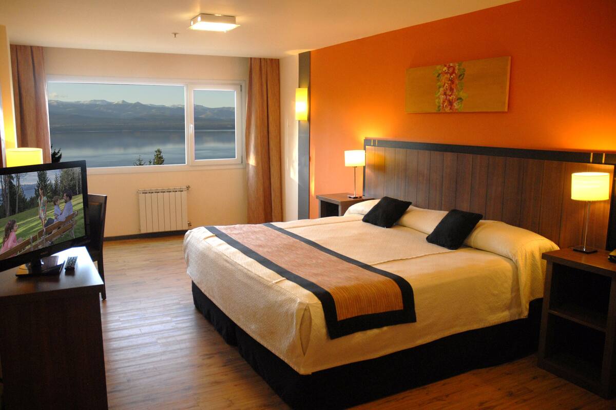 Ein Hotelzimmer mit einem großen Bett, einem Fernseher auf einem Holztisch, Blick auf Berge durch das Fenster und einem Gemälde an der Wand.