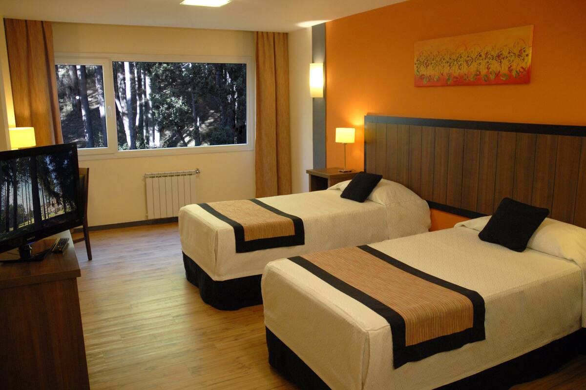 Ein Hotelzimmer mit zwei Betten, einem Fernseher, einem Fenster mit Blick auf Bäume und einem Bild an der Wand.