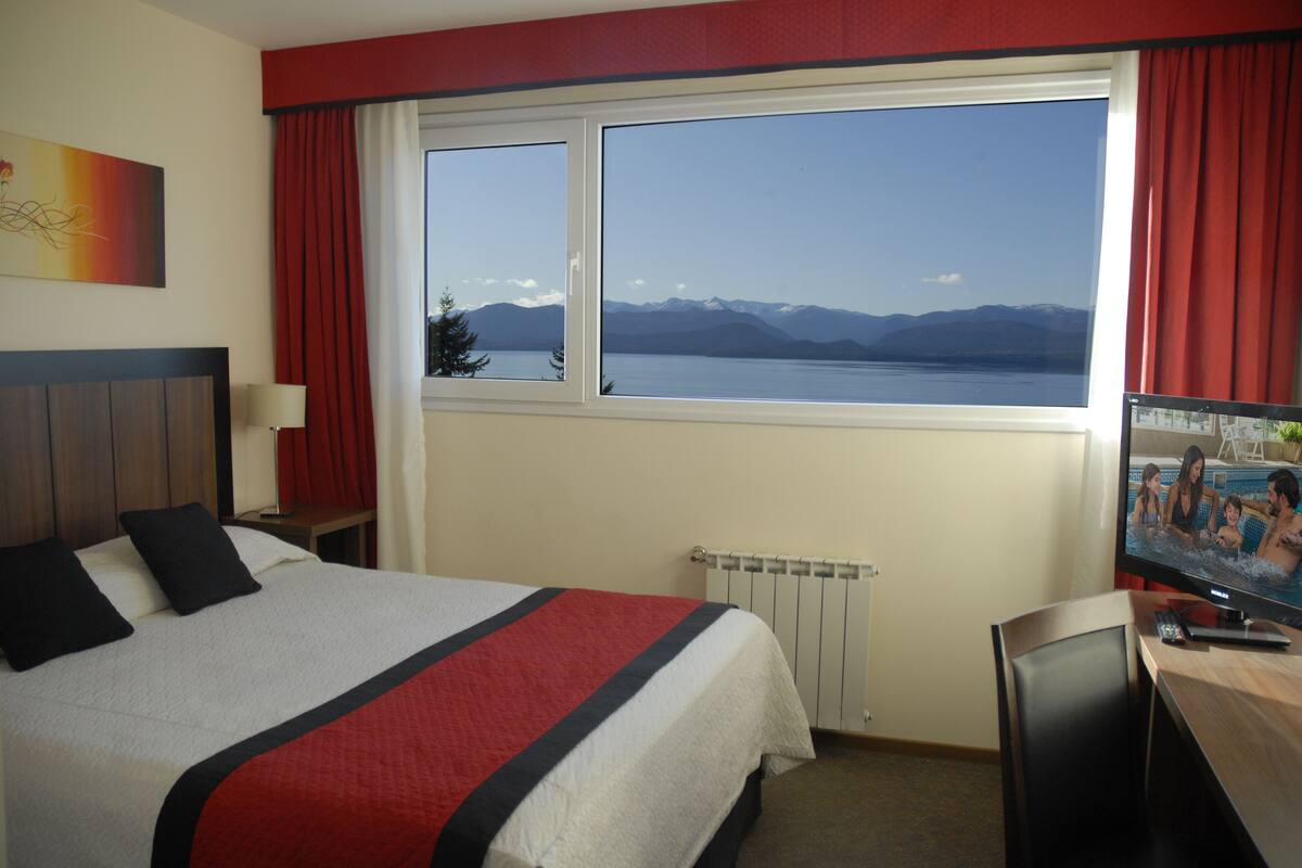 Ein Hotelzimmer mit einem großen Bett, einem Fernseher und Blick auf Berge und einen See.