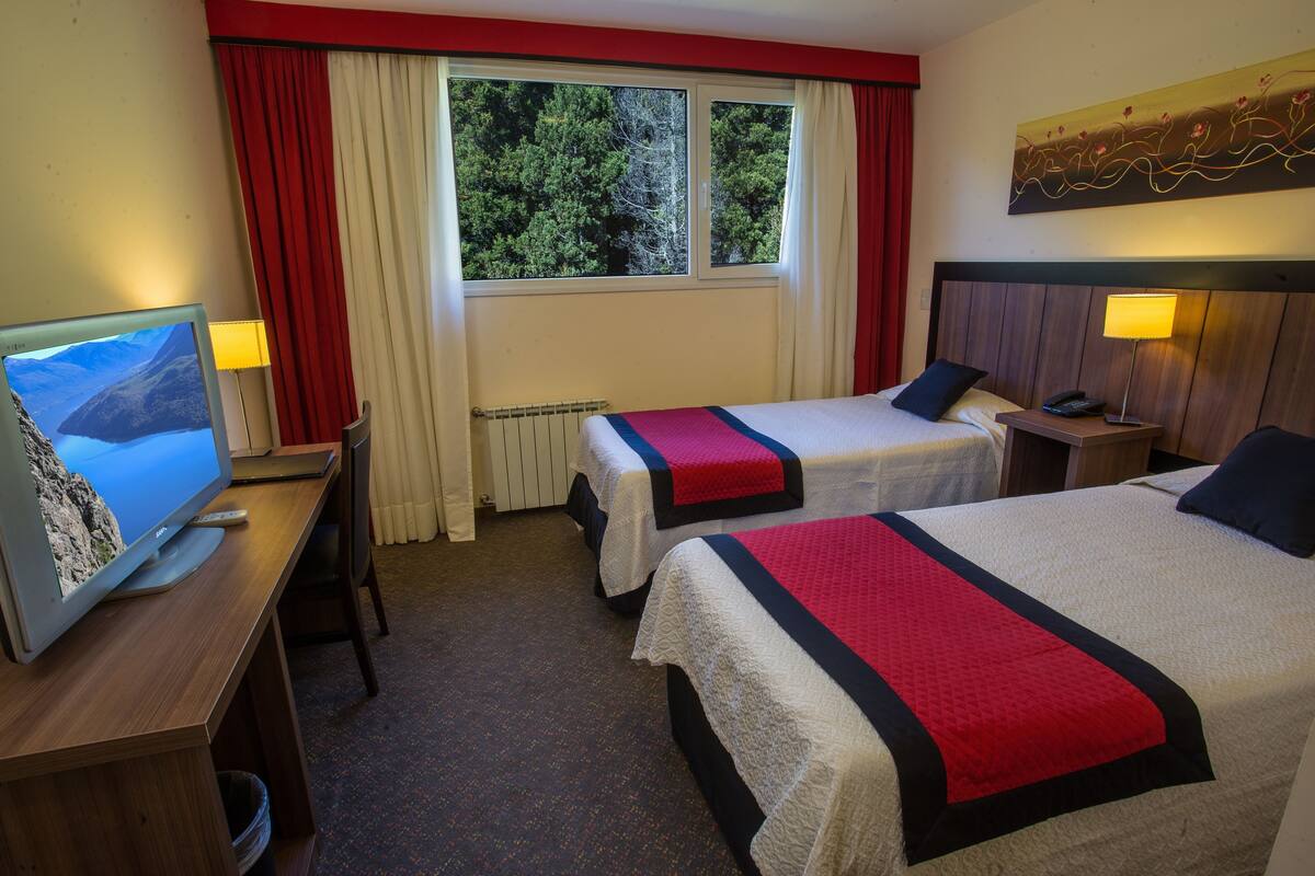 Ein Hotelzimmer mit zwei Betten, einem Fernseher, einem Schreibtisch, einem Fenster mit Vorhängen und einem Bild an der Wand.