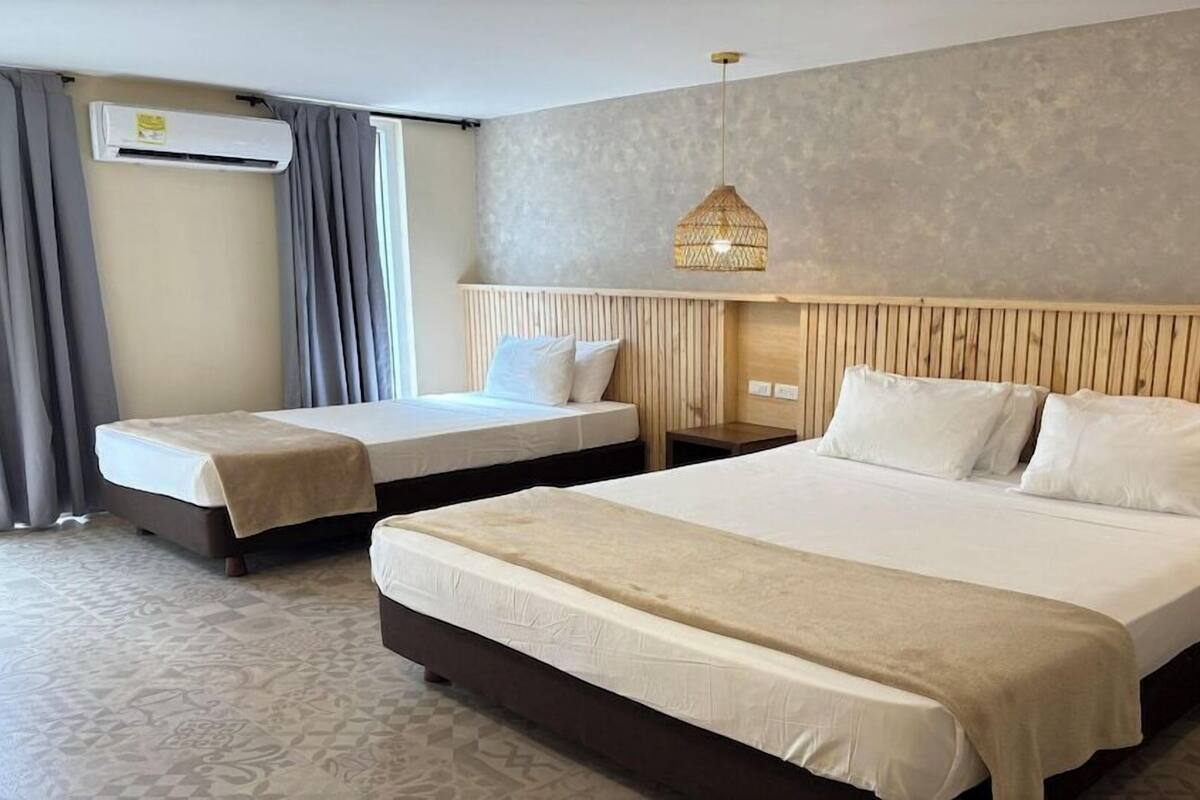 Hotelkamer met twee bedden, een groot bed in het midden en twee kleinere bedden aan weerszijden. Er is een houten hoofdbord, een wandairconditioner en een hanglamp.