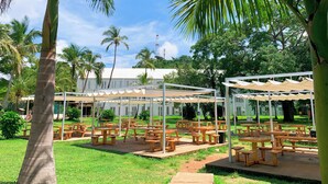 An outdoor dining area with wooden tables and benches, surrounded by palm trees and a building in the background.