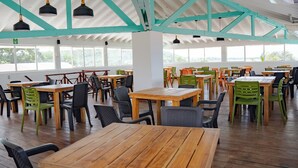 A spacious indoor dining area with wooden tables and chairs, large windows, and a high ceiling with exposed beams.