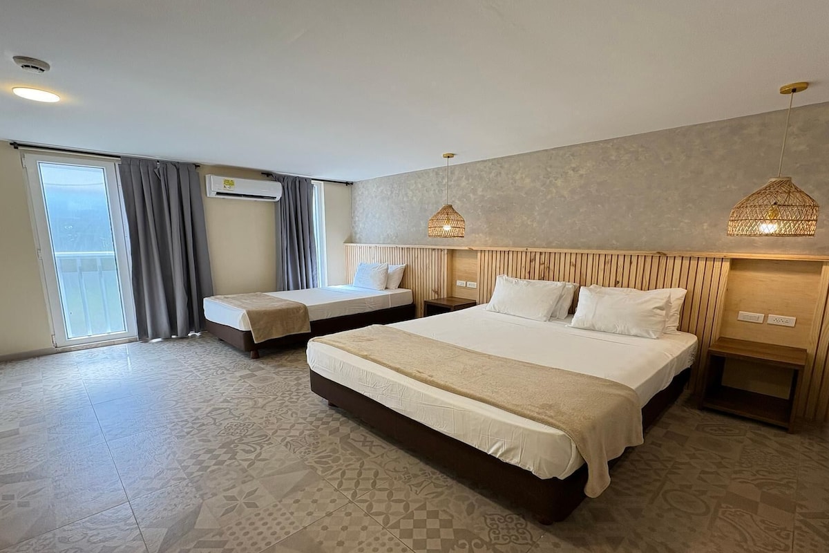 Una camera d'albergo con un letto grande, un letto più piccolo, una finestra con tende, un condizionatore d'aria a parete e un lampadario.