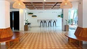 A spacious lobby with a patterned floor, wooden benches, a reception desk, and hanging plants.