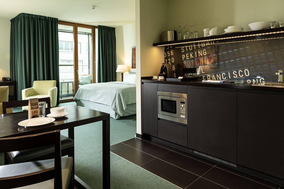 Et hotelværelse med seng, spisebord med stole, et tekøkken med mikroovn og udsigt over byen.