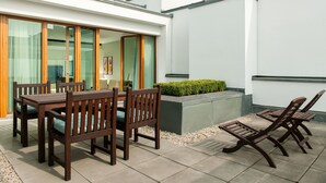 Un patio con tavolo e sedie, una piccola area giardino e un edificio con ampie finestre e facciata bianca.
