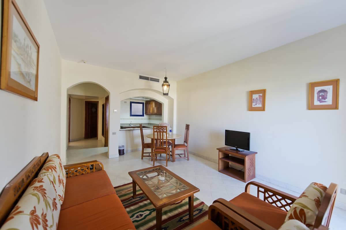 A living room with a sofa, a coffee table, and a TV stand. There is a dining area with chairs and a table in the background.