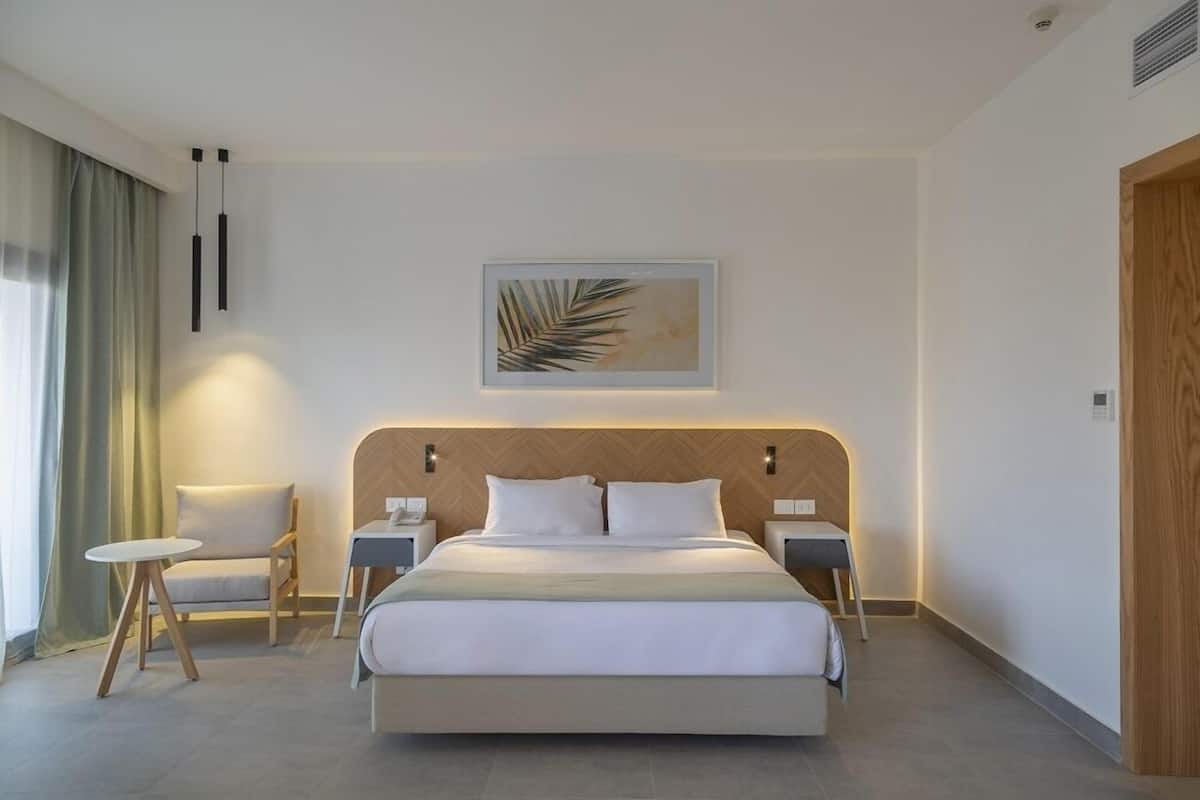 A modern hotel room with a large bed, two bedside tables, a small round table, a chair, and a wall-mounted artwork.