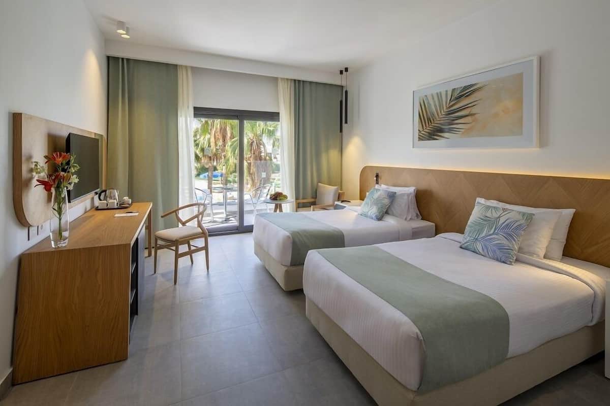 A hotel room with two beds, a desk, a chair, and a large window with a view of palm trees.
