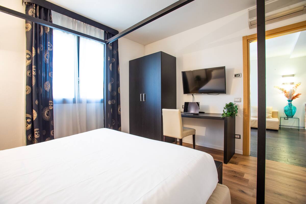 A hotel room with a bed, a desk with a computer, a TV, and a view of the hallway.