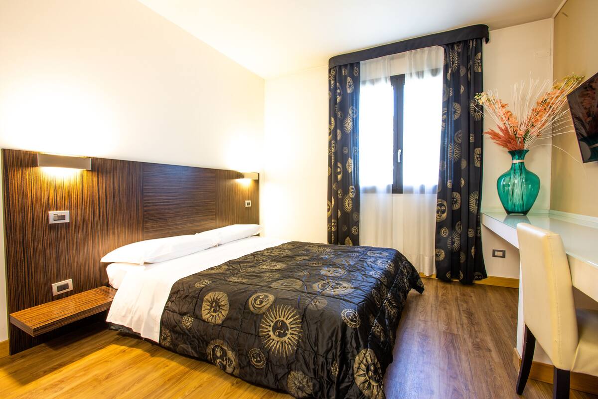 A hotel room with a wooden headboard, a bed with a patterned bedspread, a desk with a chair, and a vase with flowers on the desk.