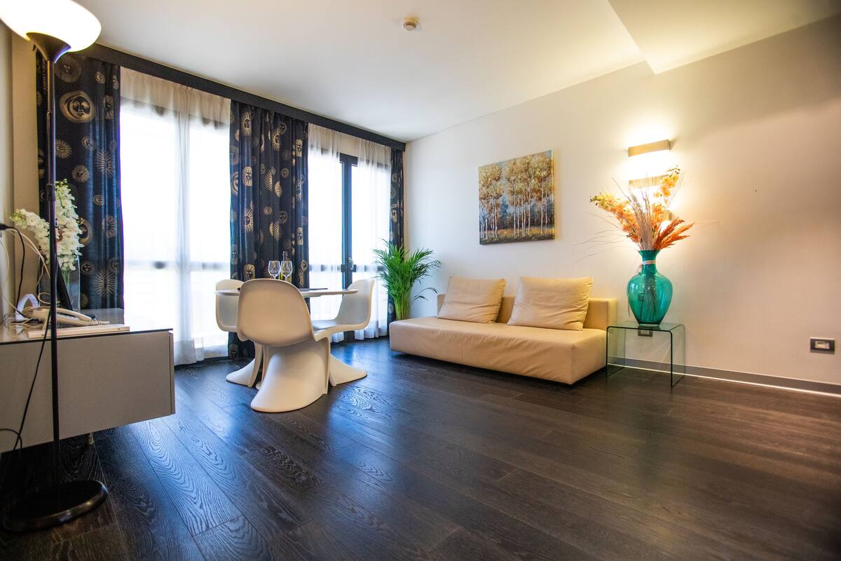 A modern living room with a sofa, a dining table, and a chair. There is a plant and a vase on a side table.