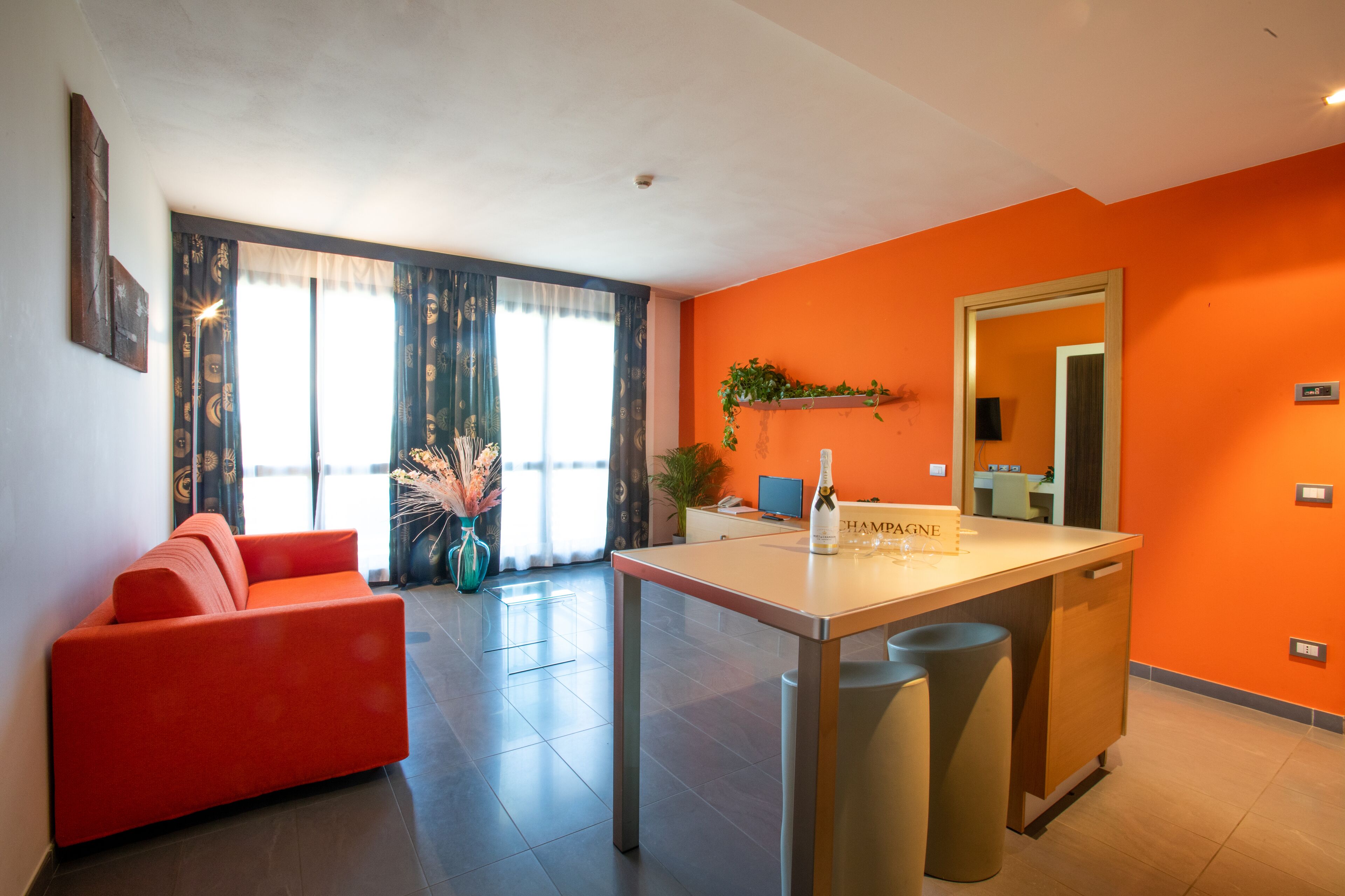 A modern living room with a red sofa, a dining area with a white table, and a kitchen area with orange walls.