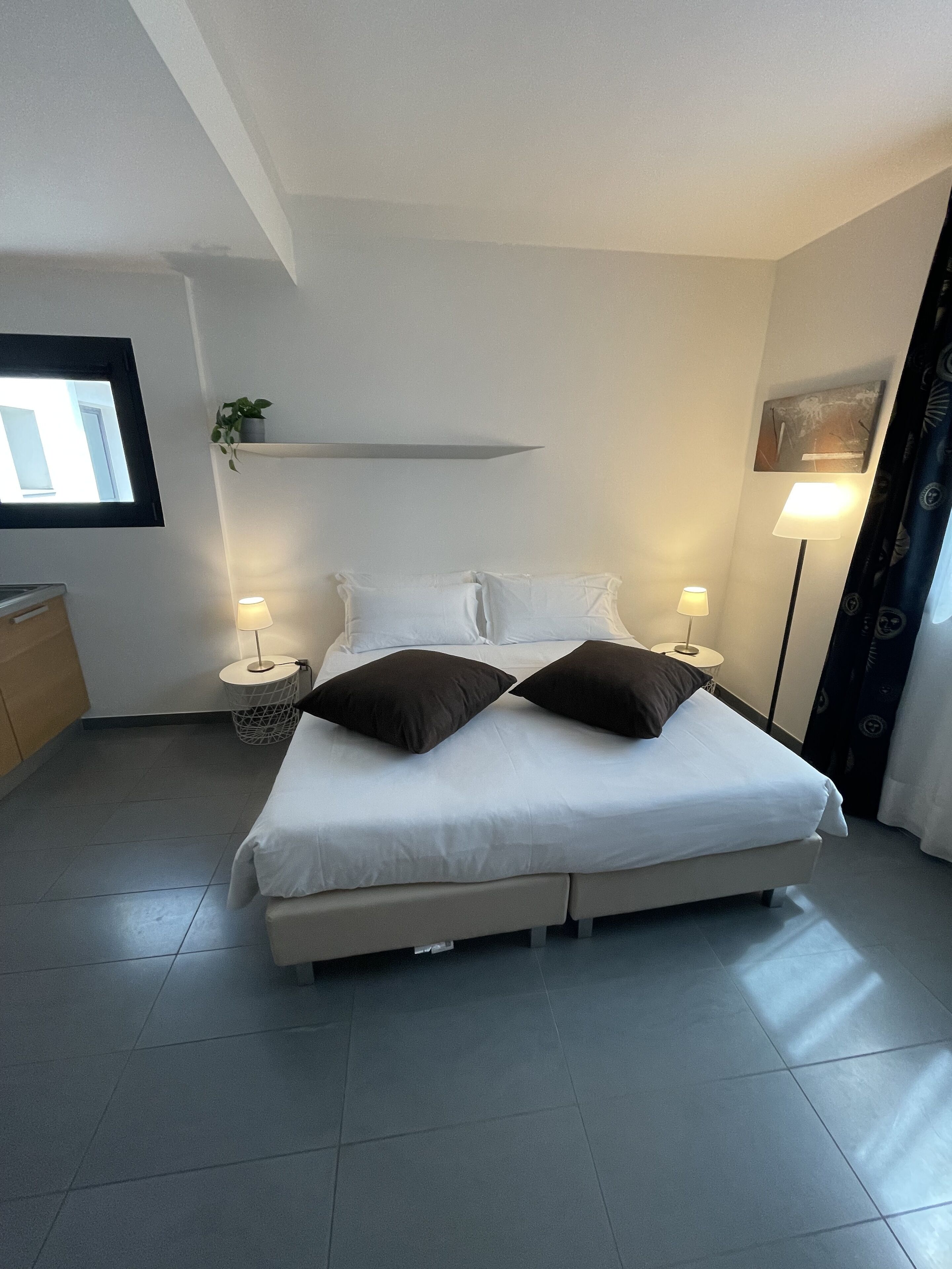 A bedroom with a bed, two bedside tables with lamps, a small shelf with a plant, a wall-mounted picture, and a window with curtains.