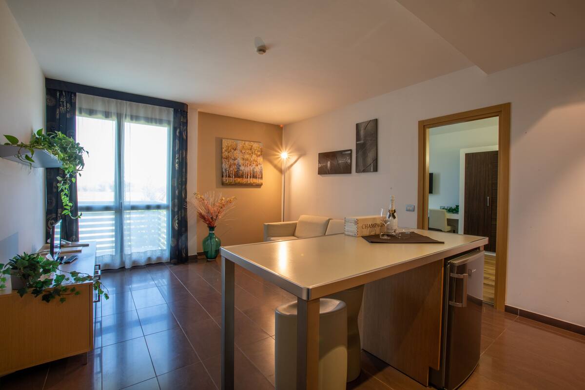 A modern kitchen with a central island, a dining area, and a view of the outdoors through large windows.