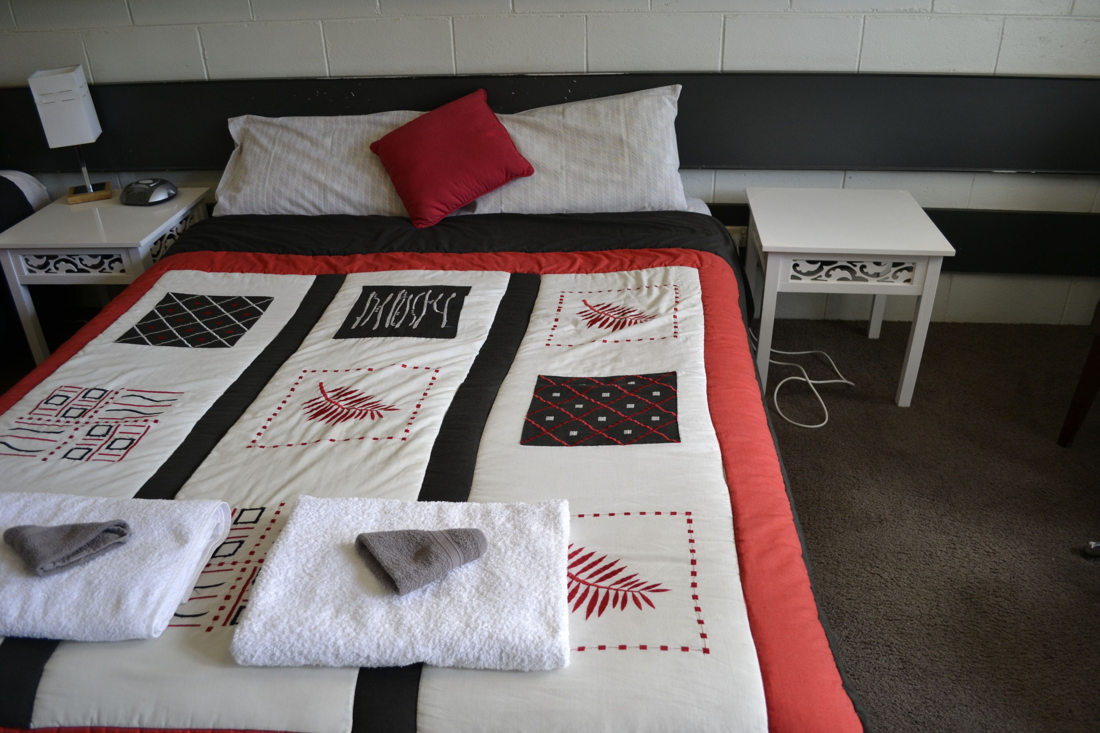 Una cama cuidadosamente tendida con un colcha roja y blanca, dos almohadas blancas y una almohada decorativa roja. Hay dos mesitas de noche blancas con diseños decorativos, una con una lámpara y un teléfono, y un cable de corriente en el suelo.