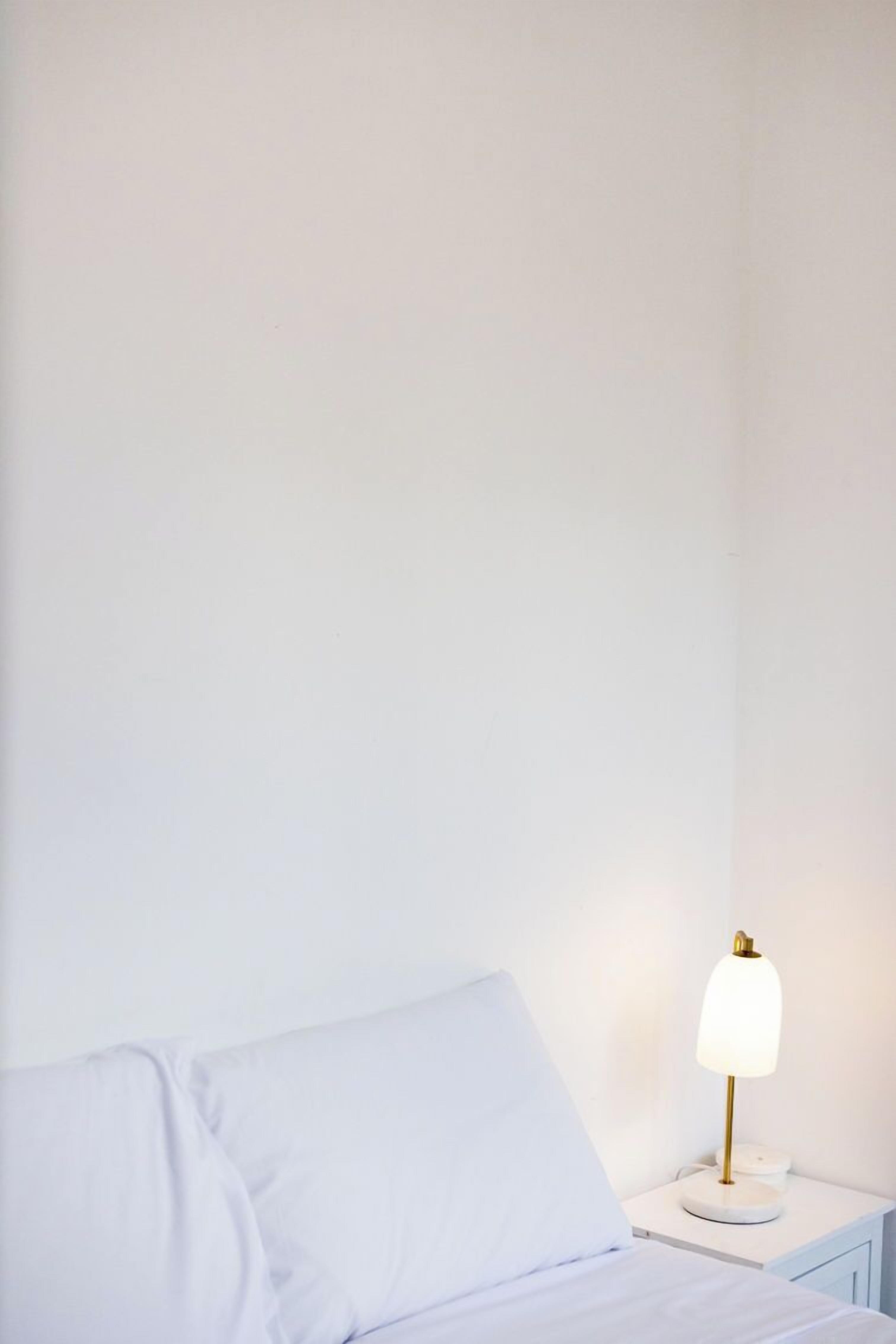 Una cama bien hecha con sábanas y almohada blancas, junto a una mesita de noche blanca con una lámpara dorada.