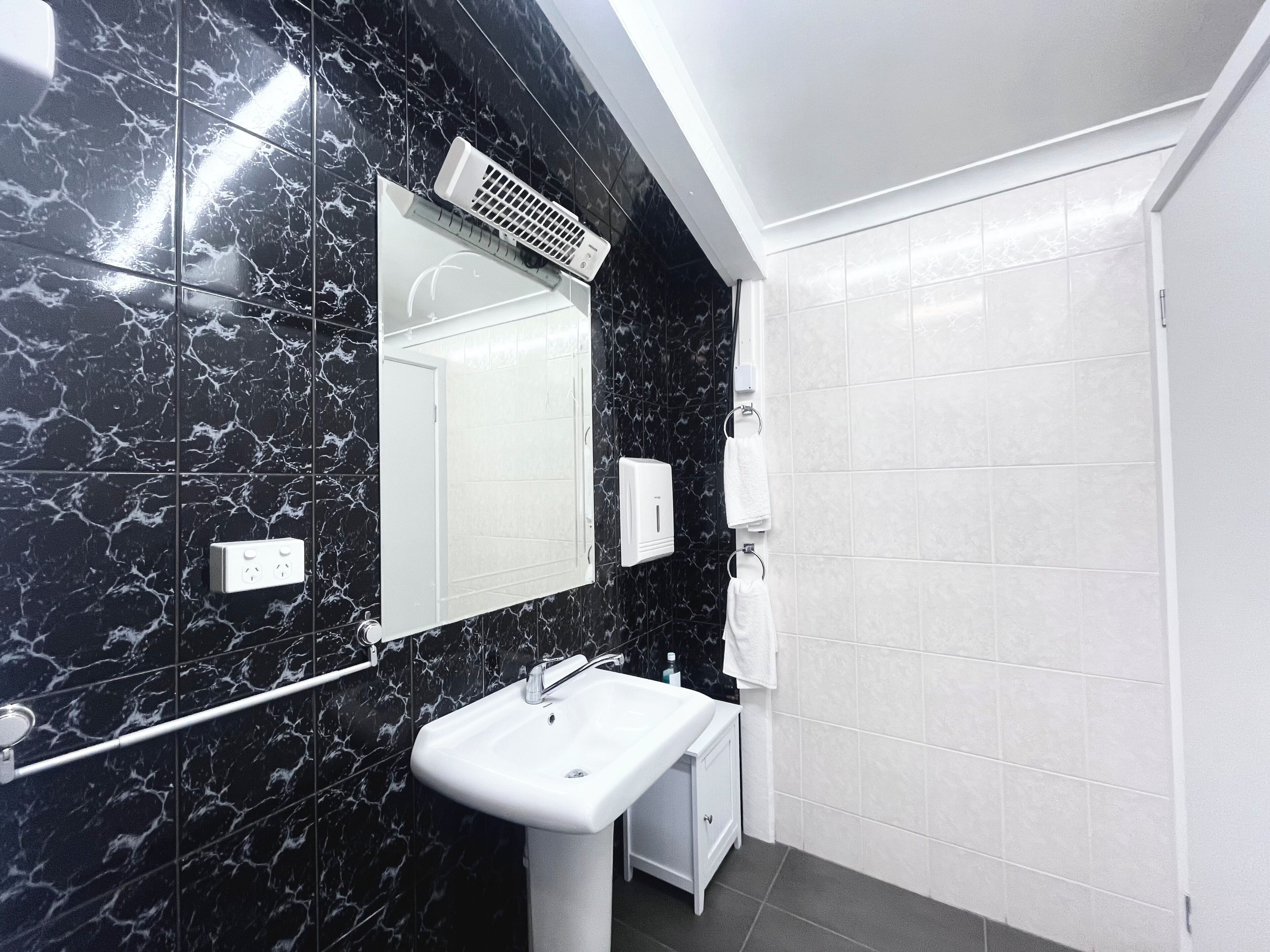 A modern bathroom with a black marble wall, a white sink, a mirror, a towel dispenser, and a shower area.