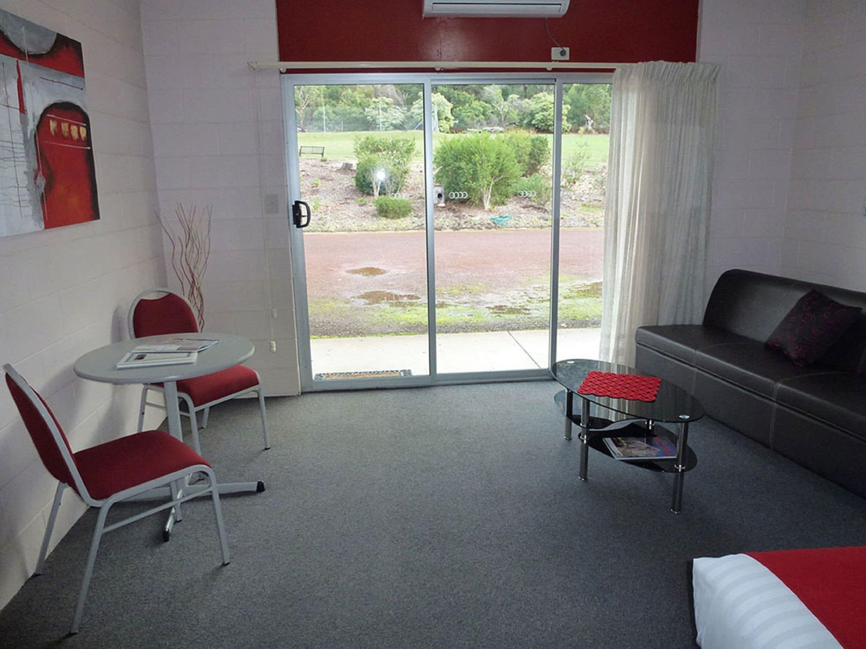 Habitación de hotel con sofá, una pequeña mesa, dos sillas, un amplio ventanal corredero que da a un espacio exterior y un equipo de aire acondicionado instalado en la pared.